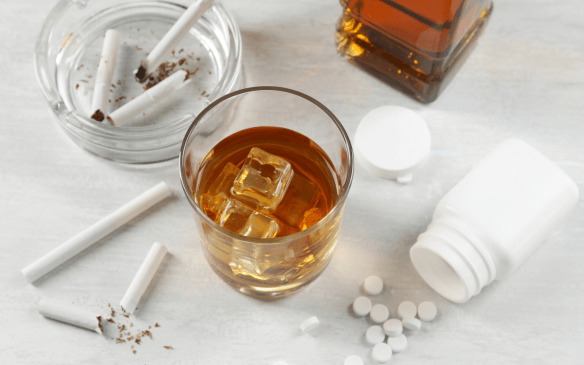 Bebida alcoólica, cigarros e comprimidos sobre uma mesa, representando abuso e dependência de substâncias.
